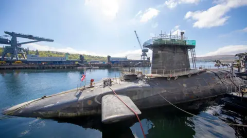 PA Media A grey submarine, partly submerged in the water, sits in a dock on a bright sunny day. The boat is covered in rigging and has a Union Jack on top of it.