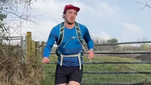 Rhodri running, with a gate and field in the background. He is wearing a long sleeved blue top, black shorts and a red baseball cap and wearing a backpack