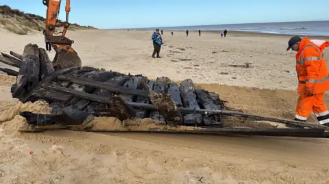 Hemsby Lifeboat The timbers from an old boat are moved up the beach. The timbers are leaving a trail in the sand. A man in hi-viz jacket and trousers is walking behind the timbers. In the background, an onlooker is standing on the beach is watching the operation.