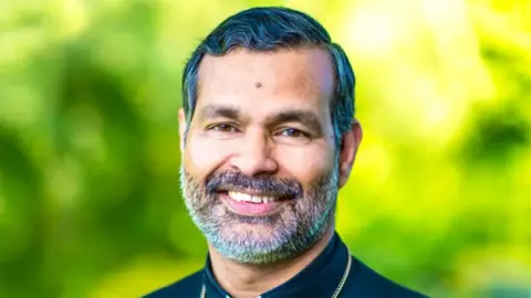 The Right Reverend Doctor John Perumbalath, with a grey beard and dark grey hair, smiles for the camera with trees in the background