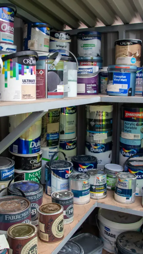 Lincolnshire County Council Lots of tins of paint of different sizes and brands on shelves inside a shed.