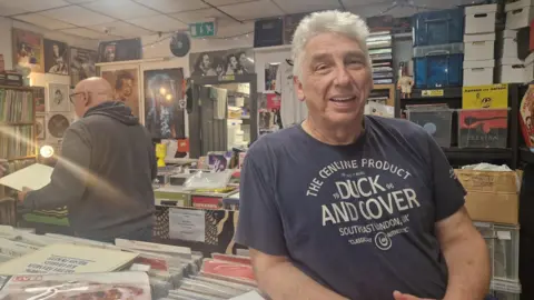 Liz Saul/BBC A man with light grey hair, wearing a faded dark blue T shirt, with large writing on it saying: DUCK AND COVER, SOUTHEAST LONDON UK. He is leaning on a row of vinyl records, which are part of several rows inside a record shop.