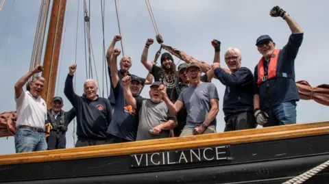 A crew of 11 men raise their hands in the air on board a boat called Vigilance.