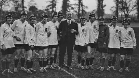 Getty/Topical Press Agency English comedian, singer and actor George Robey (1869 - 1954) with the English team, represented by Dick, Kerr Ladies from Preston, during the England vs France Women's International Football match in aid of shipwrecked mariners at Herne Hill velodrome in London, UK, 12th May 1925; embracing Robey is English winger Lily Parr and fifth from left is captain Florrie Redford. 