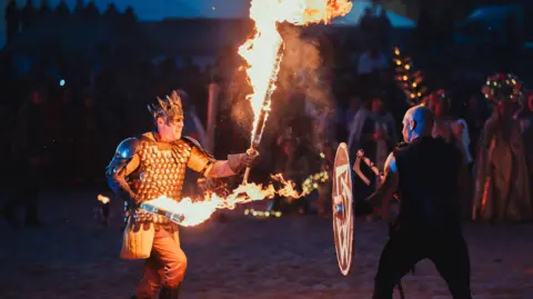 Two men in armour are performing a battle on the beach at night. The man on the left wears a crown and dis holding two flaming sword-like sticks. The man on the right holds a shield and an aze. You can see crowds watching behind them. 
