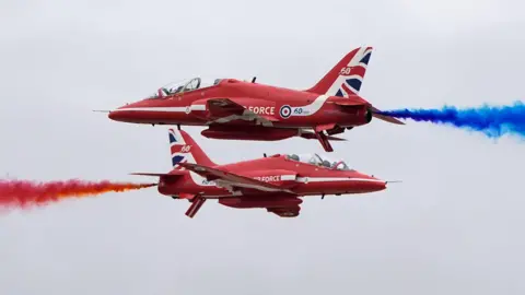 The Red Arrows display team crisscrossing, with red and blue smoke billowing from behind them.