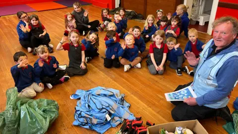 BBC/RICHARD EDWARDS A man - Mick Couzens - sits on a classroom floor, surounded by a ring of young children, and a number of adults in the back row. In front of him, there is a pile of sky-blue vests, a pile of rubbish bags and a pile of litter-grabbers with red and black handles . Couzens holds a sheet from which he appears to have been reading. Everyone is waving at the camera.