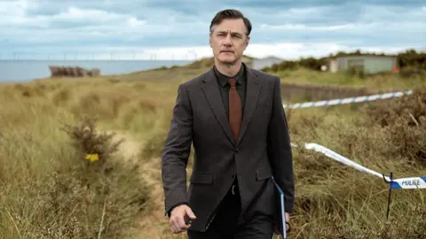 BBC David Morrissey vestindo terno atravessa o campo em cena do drama da BBC Sherwood