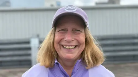 A woman with blonde hair smiling at the camera. She is wearing a lilac hat with the Brighton & Hove Albion logo on the front, and a lilac jumper
