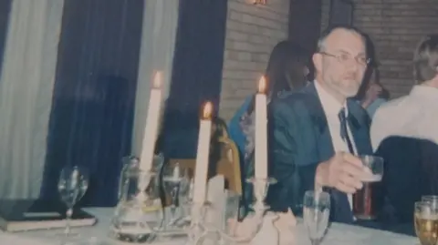 Supplied Martin Daniels wearing a suit and tie and holding a pint of beer. There are candles and glasses on the table, and brick walls and curtains are in the background.