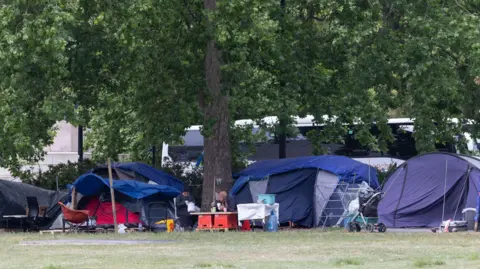 A general view of a homeless camp in Park lane. There are five tents in shot and two people with blurred faces are seated doing what appears to be eating around a table. A white coach with blacked windows is pictured in the background and in-between the tents is a tree trunk. 