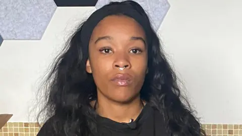 Isabel Rose is seen wearing a black top and a small nose ring, sits facing the camera indoors. A cardboard box is visible beside her and hexagonal acoustic panels are mounted on the wall behind her.