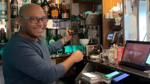 A man smiling whilst making a coffee behind a check in desk at his restaurant Cafe Spice. The glass wall behind has shelves of different alcohols.
