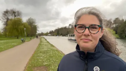 Polly has grey hair in a pony tail and wears glasses with multi-coloured rims. She wears a blue fleece and is standing by the river.