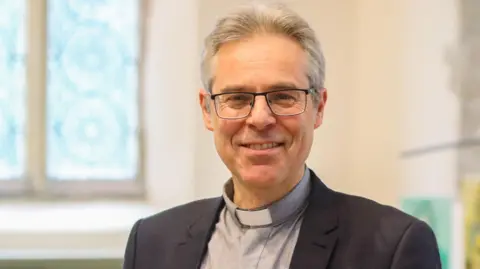 Bishop Designate Rick Simpson is smiling while stood inside a church building with stained glass behind him. He has medium length grey hair and has a clerical collar on and a suit jacket.