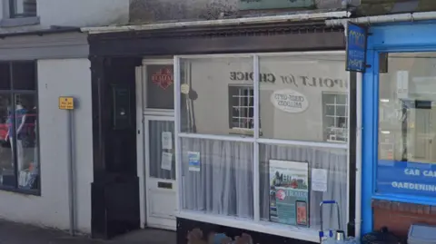 The front of a premises on a street with a large square window, half-covered by net curtains