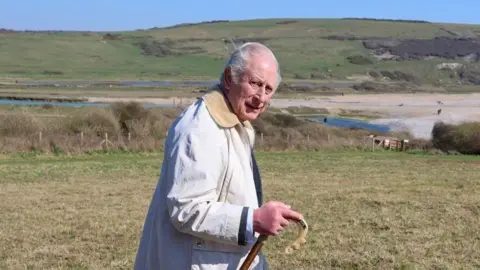 King Charles III looks in the direction of the camera as he walks. He is wearing a white coat and carrying a wooden walking stick. Behind him there is a beachy area, and beyond that a hill. 
