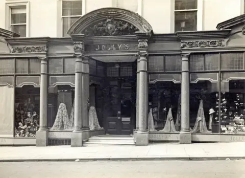 An archive picture of the exterior of Jolly's taken from the street. It has columns and large windows, with draped fabrics displayed in the front. The picture is in black and white, and was taken in the 1930s. 