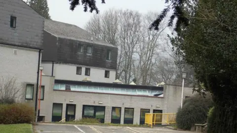Facebook A general view of the assessment centre, which is grey brick with a row of ground floor windows and a row of skylight windows above. A concrete car park can be seen in front of the building with a yellow railing on the right hand side. 