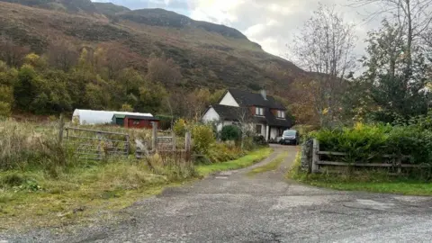 BBC A house is at the end of a track. There's a minibus parked outside. There are sheds next to the house, a plastic gardening tunnel and a hill in the background.