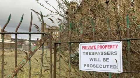 Charles Heslett/BBC A bent metal sign attached to green, rusting railings which reads 'Private Property' in red lettering and then 'Trespassers will be prosecuted' in black underneath with part of the mill complex visible in the distance.