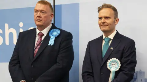 PA Media Two men in suits and rosettes stand side by side, the man on the right looks happier.