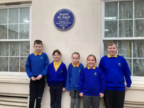 Five primary-aged children in blue school uniforms and yellow polo shirts stand below a sign that reads: Guernsey Museums - Jessie de Garis 1916-1984 Pioneer of Special Education taught here.