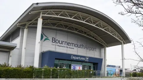 Bournemouth Airport Departure building - a grey building with a large portico held up by a number of columns and a blue surrounding to the glass entrance and a green hedge at the front.