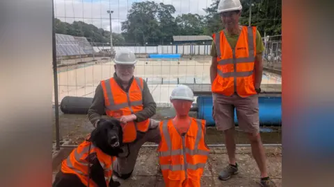 Laura Alliston Two men, a dog, and for some reason a crash test dummy, are all donning orange high-visibility jackets. The men are wearing hard hats. The dog doesn't get one. Behind them, surrounded by construction fencing, is the open-air pool - completely dry and empty.