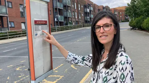 A woman, Nursen Smith, wearing a white top with black patterns on it. She is pointing at a bus timetable on a post.
