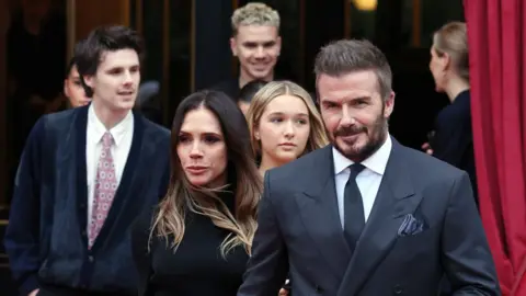 Getty Images Lady Beckham and Sir David Beckham and family leaving their hotel during Haute Couture Spring Summer 2026 as part of Paris Fashion Week on January 26, 2026 in Paris, France. Sir David is slightly in front in a suit and tie, with Lady Beckham behind him in black with her hair down
