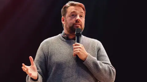 Jason Manford wearing a grey jumper over a shirt. He is holding a microphone and talking into it while expressing himself with his hands.