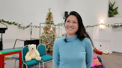 A woman with black hair and stood in a room which has Christmas decorations all around and an array of toys behind her.