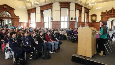 BBC Rows of teenagers wearing scout neckerchiefs sitting in a wood-panelled room. At the front of the room, a woman in a polo shirt and a neckerchief is speaking to them through a microphone