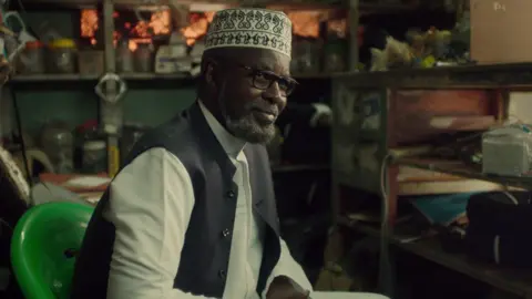 Netflix Ahmed Five sitting down on a gren plastic chair in a workshop wearing a traditional cap and waistcoat over a white shirt.