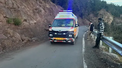 Gambar ambulans lewat saat dua pria berdiri di dekatnya 