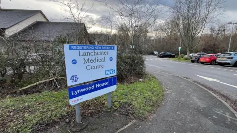 A sign reading Lanchester Medical Centre on the side of a pavement leading to a car park.
