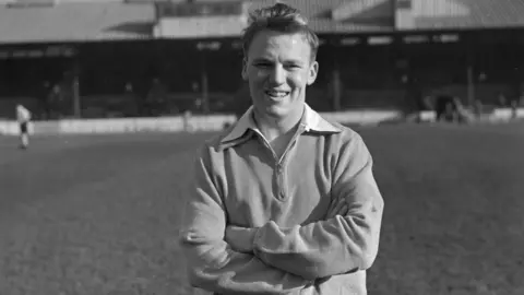 Getty Images A black and white picture of Frank Blunstone during his playing career - he is standing on a pitch with his arms folded. He has short hair and is wearing a long-sleeved top.