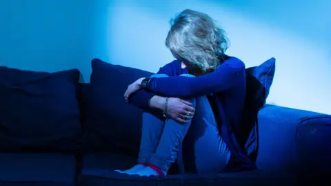 Generic image showing an unidentifiable woman with her head bowed down and hugging her knees while sitting on a blue sofa.