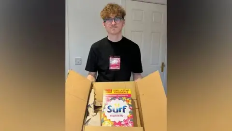 Andrea Carnell Devon stands holding a large open cardboard box which contains a large box of Surf washing powder. The box is pink and white and says "Long lasting fragrance" at the top. Devon wears glasses and has short curly blonde hair and stubble. He wears a black T-Shirt.