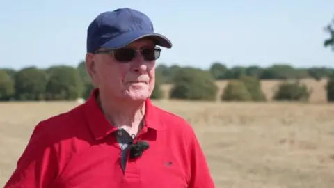 BBC A man wearing black sunglasses and a navy cap is wearing a red polo shirt and looking to the side with a serious expression. The background is blurred but you can make out fields of dry pasture and some trees.