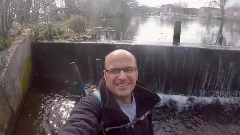 Gavin Cox A selfie of Gavin Cox smiling at the camera, which is pointing from above. He stands in front of a river weir in a waterproof jacket with a backpack on with tools in the back.