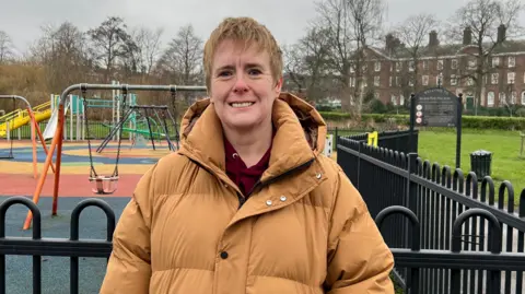 Jessica Bayley/BBC A woman with a blonde pixie cut wearing a dark yellow puffer coat stands in front of a railing. Behind her you can see a playground featuring a couple of swing sets and a slide in the background.