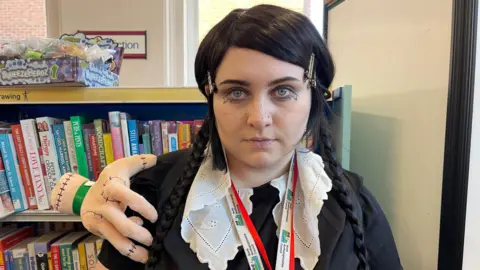 Evangeline Maddams dressed as Wednesday Addams with a black wig with two long plaits, a black dress and white collar. 