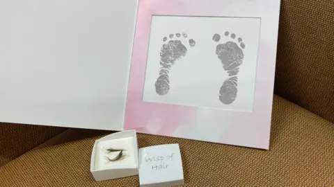 A picture of two grey small baby footprints are seen in a card pink-framed card.  A box with two small pieces of hair is open and on the lid read 'wisp of hair'.