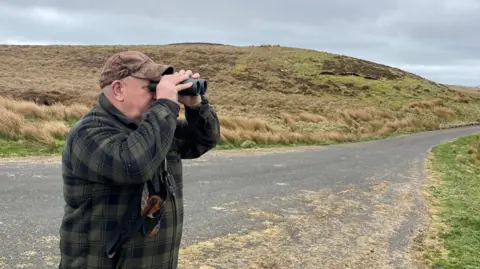 Ian Glendinning is holding binoculars to his eyes and staring across an empty moorland landscape. He is wearing a green and black check jacket and brown peaked cap.