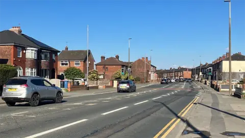Liverpool Road in Royton, Oldham, the hoped location of the scheme. It is a sunny day with a blue sky. There are several cars travelling on the left side of the road.