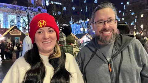 Ashley Wilie has long black hair and is wearing a cream jumper with red hat. Her partner beside her has long black and grey hair, and beard. He is wearing a grey jumper and top.