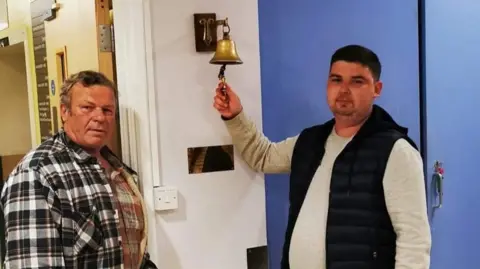 Brain Tumour Research Liam Harvey wearing a grey sweatshirt underneath a black gilet. He is standing in hospital alongside his father, who is wearing a black and white plaid jacket. Liam is ringing a gold bell which marks the end of his treatment.