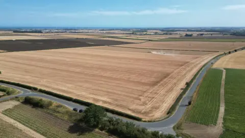 A drone shot of fields and a road, the fields are being harvested and wind turbines are visible in the far distance. 